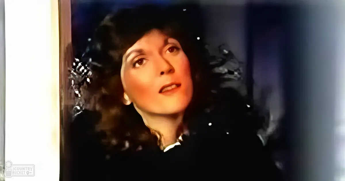 Adult female wearing a black, fitted coat with high collar and embellishments in an indoor setting with natural light.