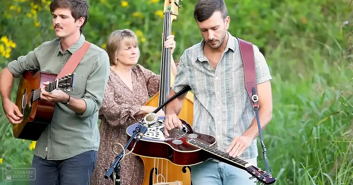 Outdoor musical performance with young adult male singer wearing a green button-up shirt, middle-aged female musician playing double bass in brown floral dress, and another young adult male holding an acoustic guitar amidst spring or summer foliage.