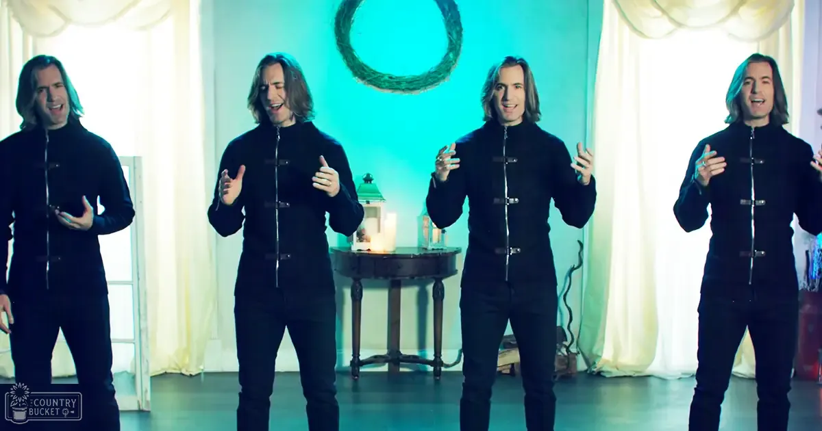 Young adult males in a staged indoor setting, possibly for a performance or promotional video; light-colored curtains, teal accent wall with circular wreath decoration.