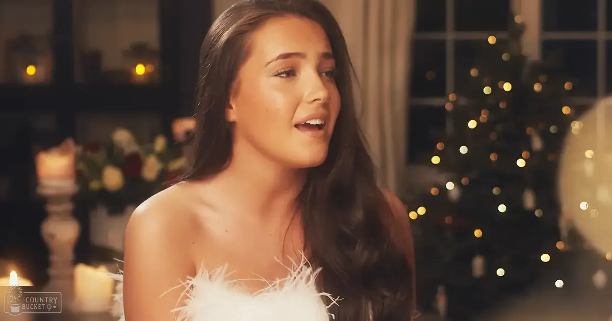 A young adult female wearing an elegant white strapless garment adorned with feather-like details at the neckline, set against a cozy Christmas-themed interior decorated for the holiday season.