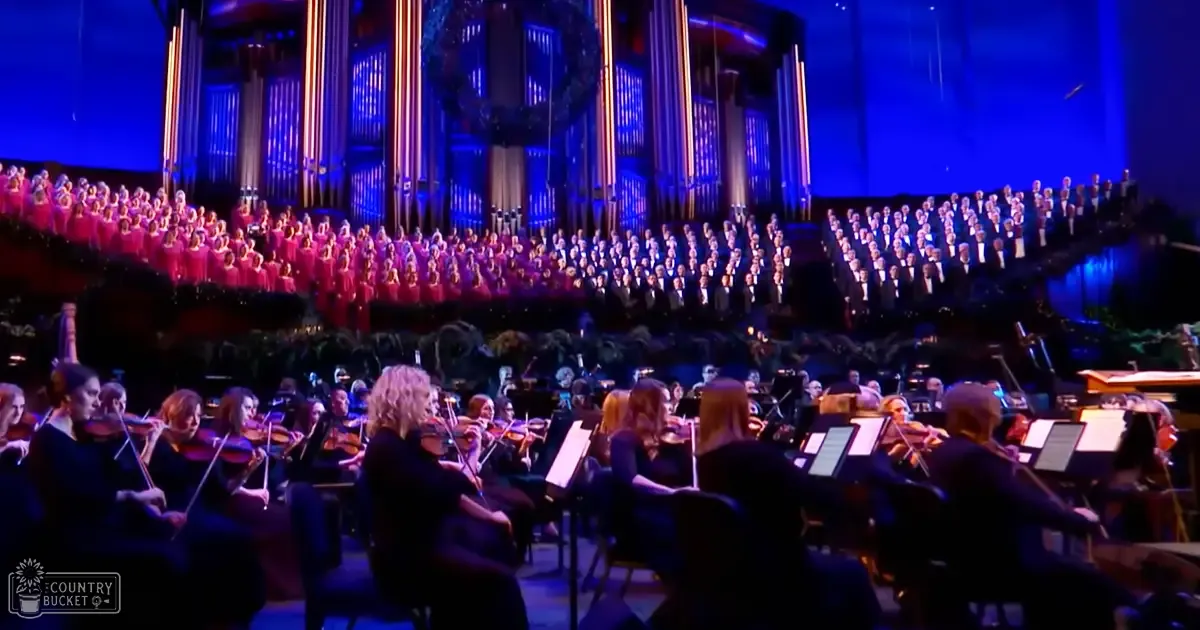A large choir and orchestra performing in an indoor auditorium with stage lighting, pink dresses on adult females, dark formal attire on adults of various ages playing string instruments.