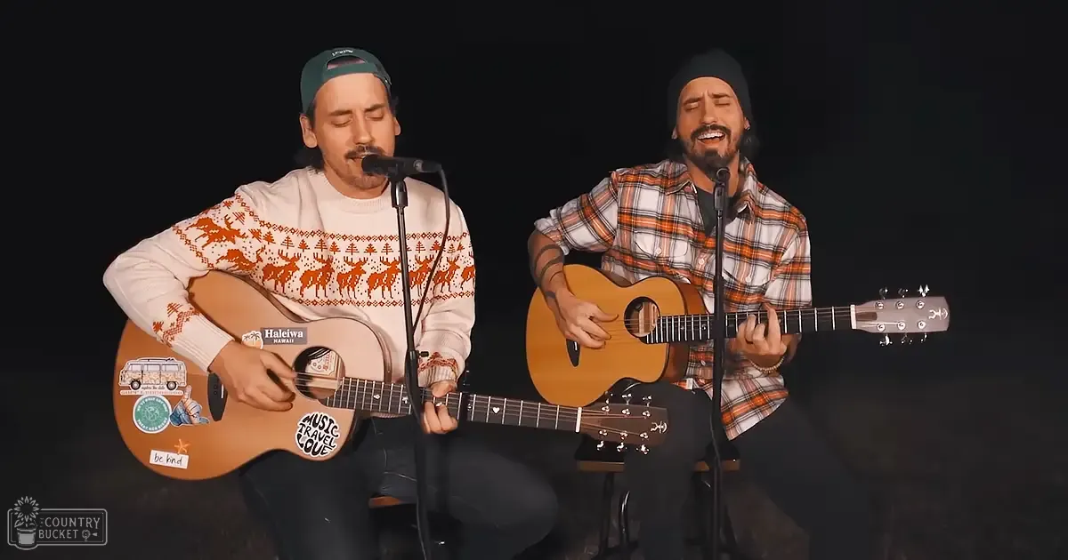 Young adult male singer in a light-colored sweater with orange reindeer and tree patterns, playing an acoustic guitar; another young adult male singer wearing an orange and white plaid shirt, holding a guitar. Both are performing indoors against a black background.