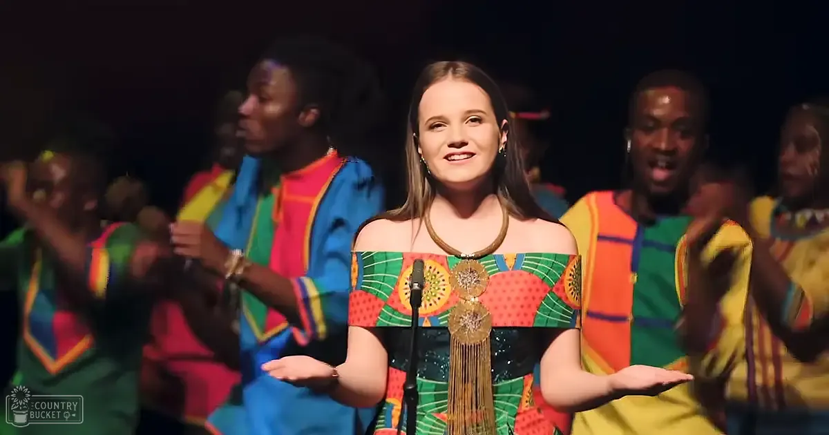 A young adult female in a flowy, off-the-shoulder dress with fruit or floral print stands in the foreground of an indoor event or performance. Multiple individuals in matching brightly colored striped shirts are part of what appears to be a choir or group performance.