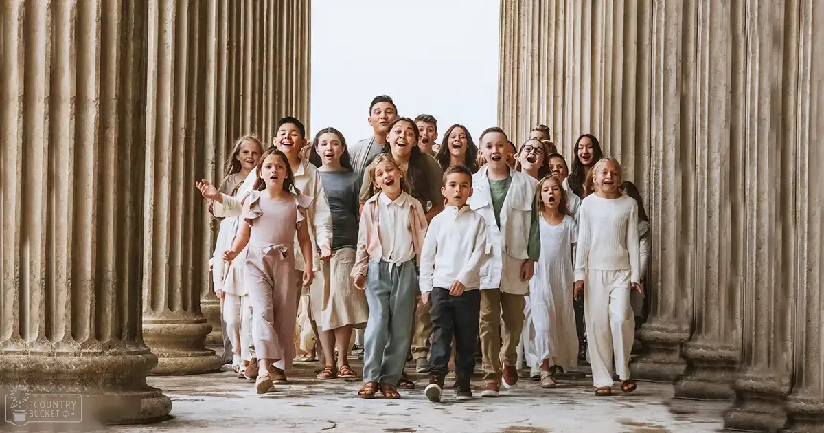 Children in an indoor setting with large columns, wearing light pink ruffled tops, white dresses, and various casual outfits. The scene suggests a historical or architectural space like a temple.