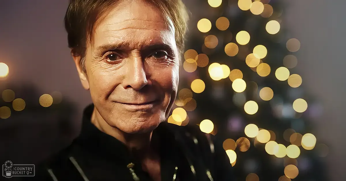 An older adult male wearing a dark, textured jacket or sweater with zippers, set against festive bokeh lights suggesting Christmas time.