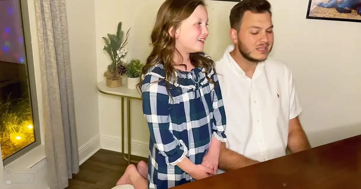 A young girl in a blue and white plaid dress stands indoors with an adult male wearing a white button-up shirt, surrounded by holiday decorations.