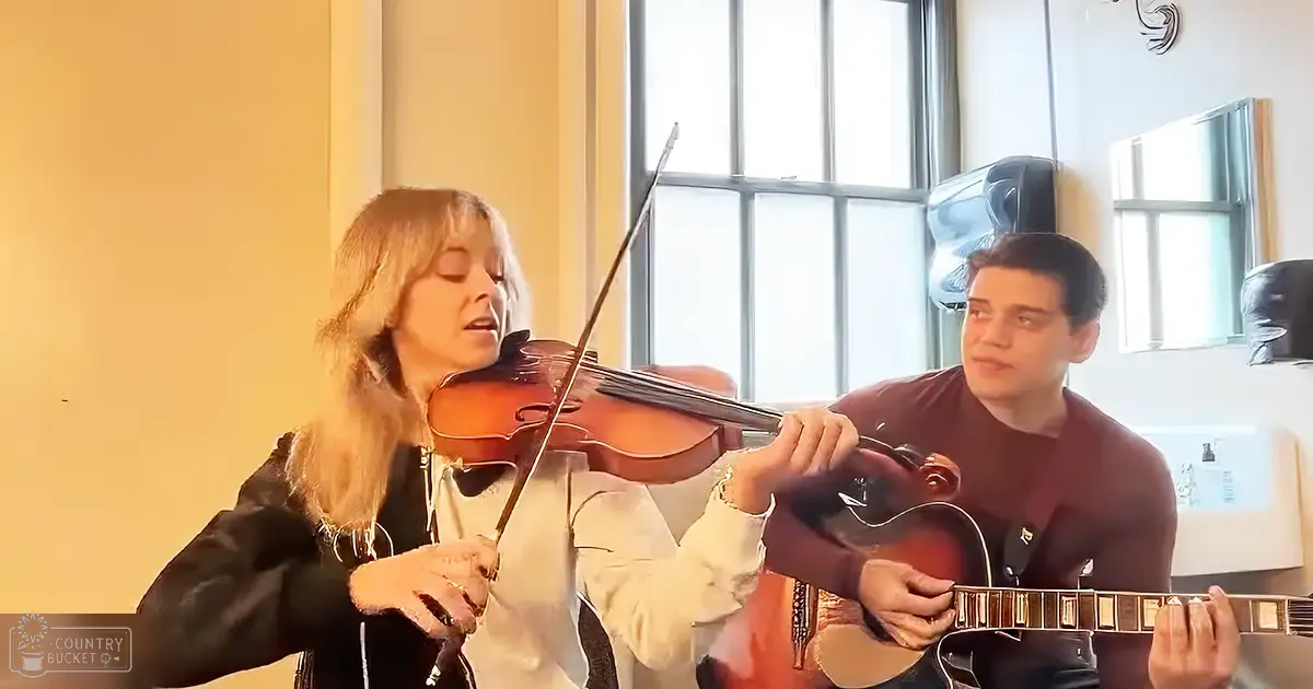 A musical performance with two musicians playing inside, natural light streaming through large windows, beige walls, white window frame, brown guitar and shirt.
