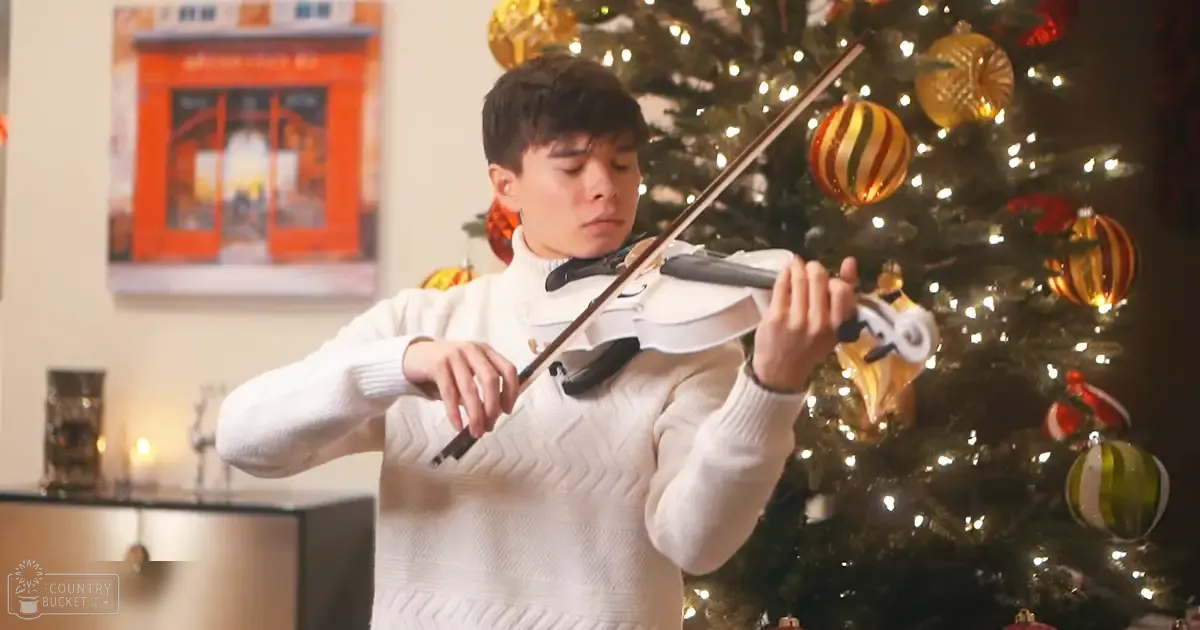 A young adult male plays the violin in front of a decorated Christmas tree, wearing a white sweater with textured pattern and ribbed cuffs. The scene is set indoors during the holiday season.