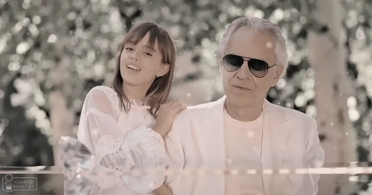 A young girl wearing a white lace dress and an older adult male dressed in a white blazer, set against lush greenery with soft diffused light suggesting spring or summer.