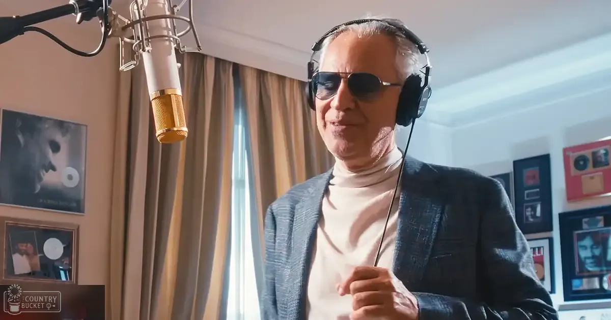 Older adult male in a recording studio, wearing dark blazer over light turtleneck sweater with textured pattern, surrounded by professional microphone and framed awards, certificates, photographs related to music or recording.