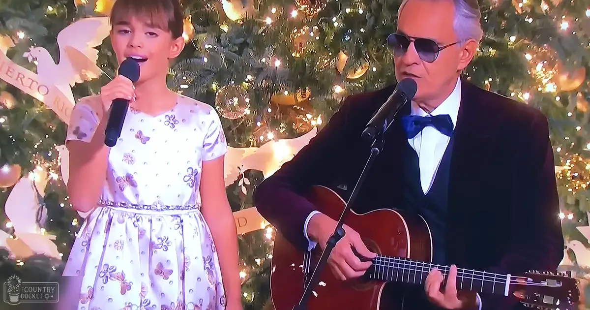 A child singer in a white dress with purple butterfly patterns and an older adult male guitarist in formal attire performing indoors during the holiday season, with a decorated Christmas tree in the background.
