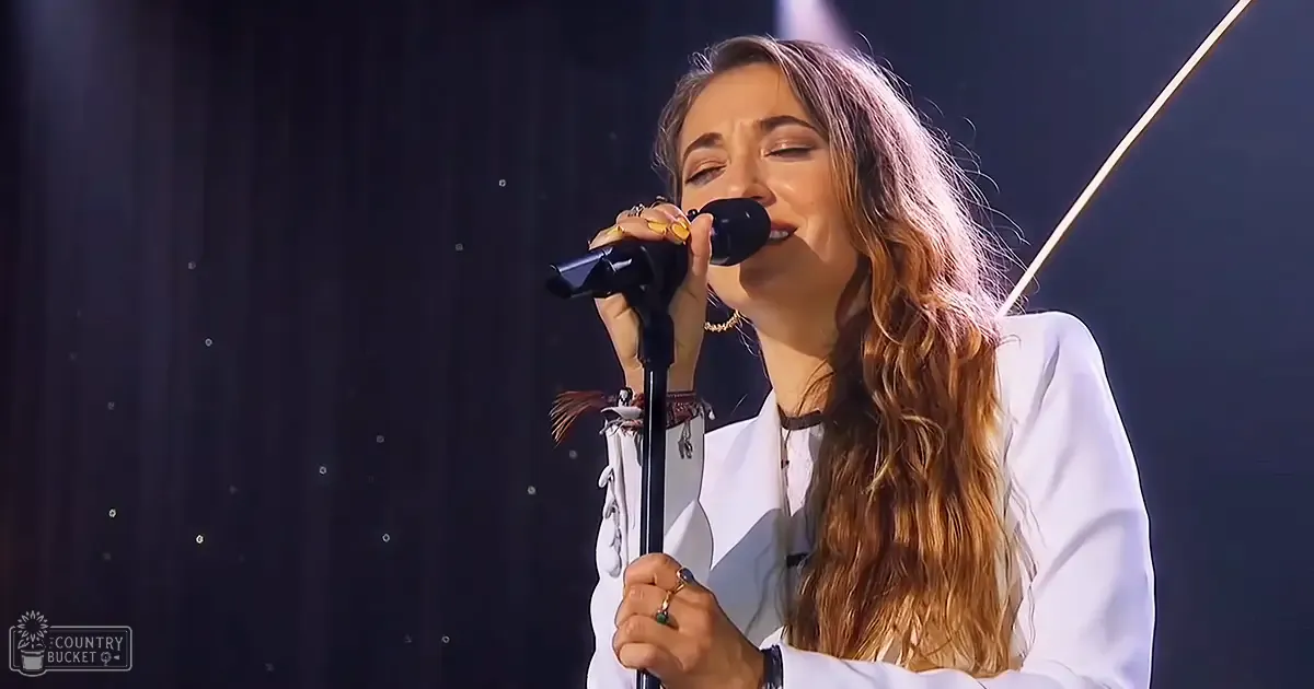 A young adult female wearing a white, loose-fitting blouse with long sleeves performs on stage under spotlights. She wears multiple rings and bracelets.