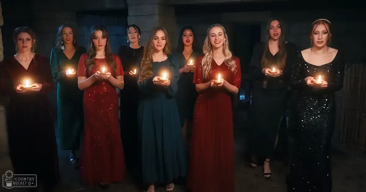 A group of women dressed in vintage 1950s-style dresses stand together inside what appears to be a church with stone walls and pillars. Dim lighting is provided by candles held by the individuals.