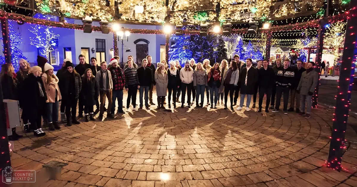 A festive outdoor scene with a group of teens/young adults dressed in winter attire, decorated for Christmas. String lights and holiday decorations illuminate the setting at night.