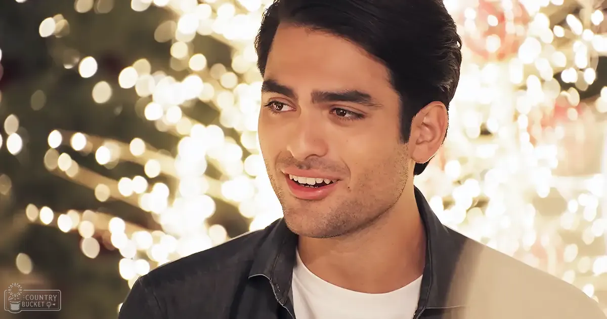 A young adult male in a relaxed indoor setting with festive holiday lighting, wearing a dark jacket over a white t-shirt.