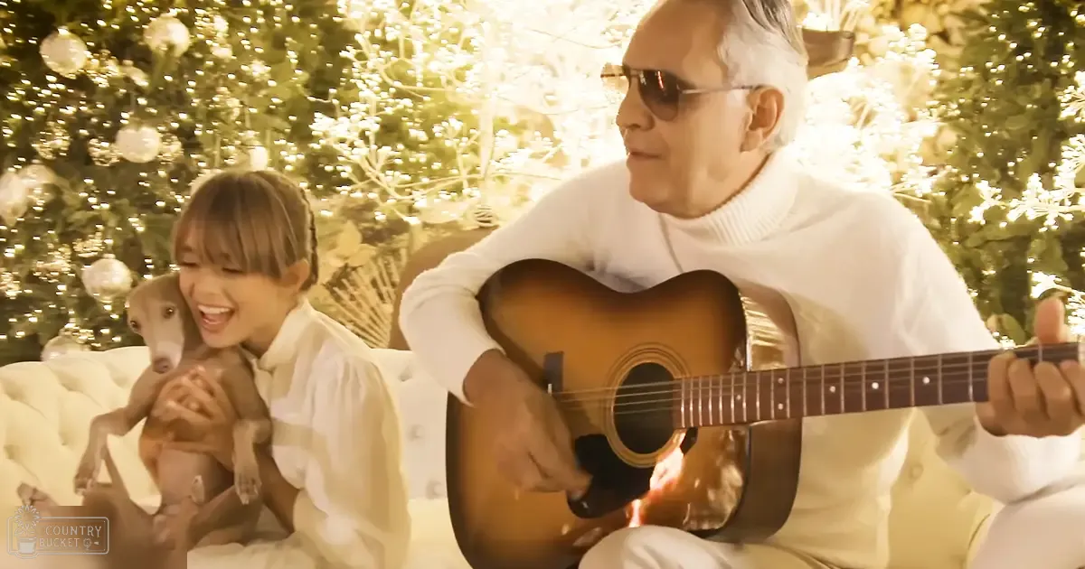 An older adult male singer playing guitar indoors with a Christmas tree and a whippet dog, cozy indoor setting, warm golden lighting, white couch, holiday season