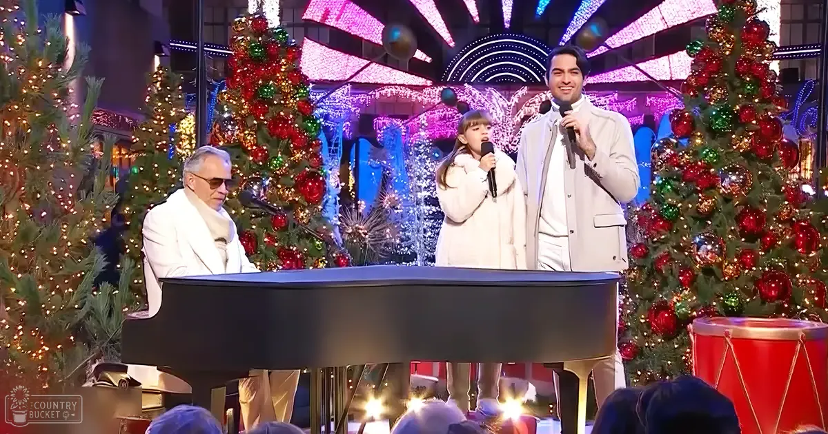 A festive holiday performance scene with an adult male singer, a child holding a microphone, and another adult male, all dressed in white coats with high collars, set against Christmas trees decorated with red, green, and gold ornaments under artificial evening lighting.