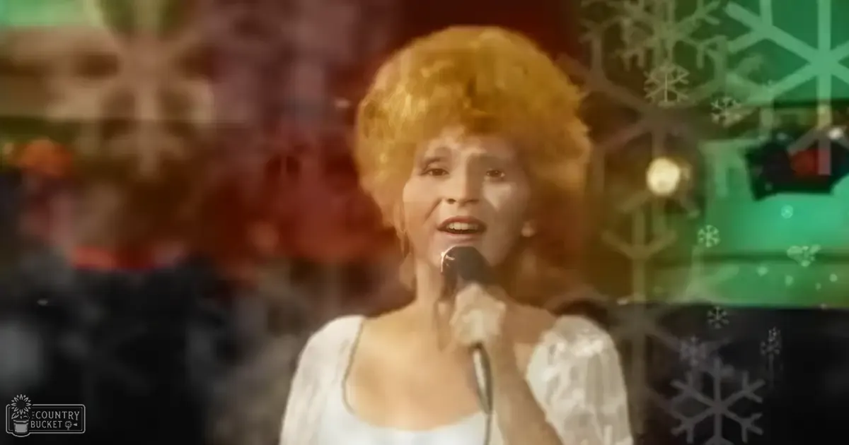 A female performer wearing a white, mid-20th-century cotton or linen dress with puffed sleeves sings into a microphone against a winter-themed studio backdrop adorned with snowflake decorations.