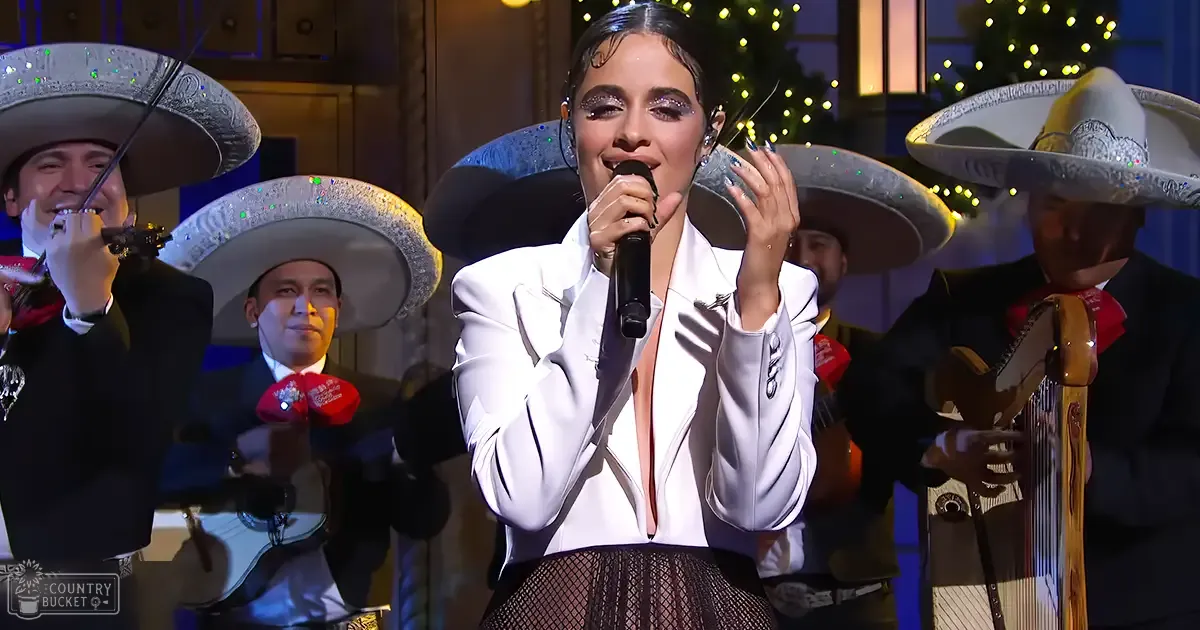 A young adult female singer performing in front of a mariachi band during what seems to be an indoor holiday event with Christmas lights and festive ambiance.