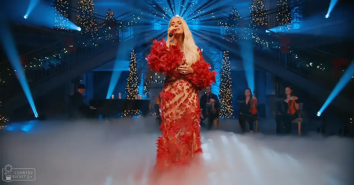 A female performer in a red floral dress holding a microphone stands on stage with other musicians playing stringed instruments, surrounded by holiday decorations under dramatic lighting.