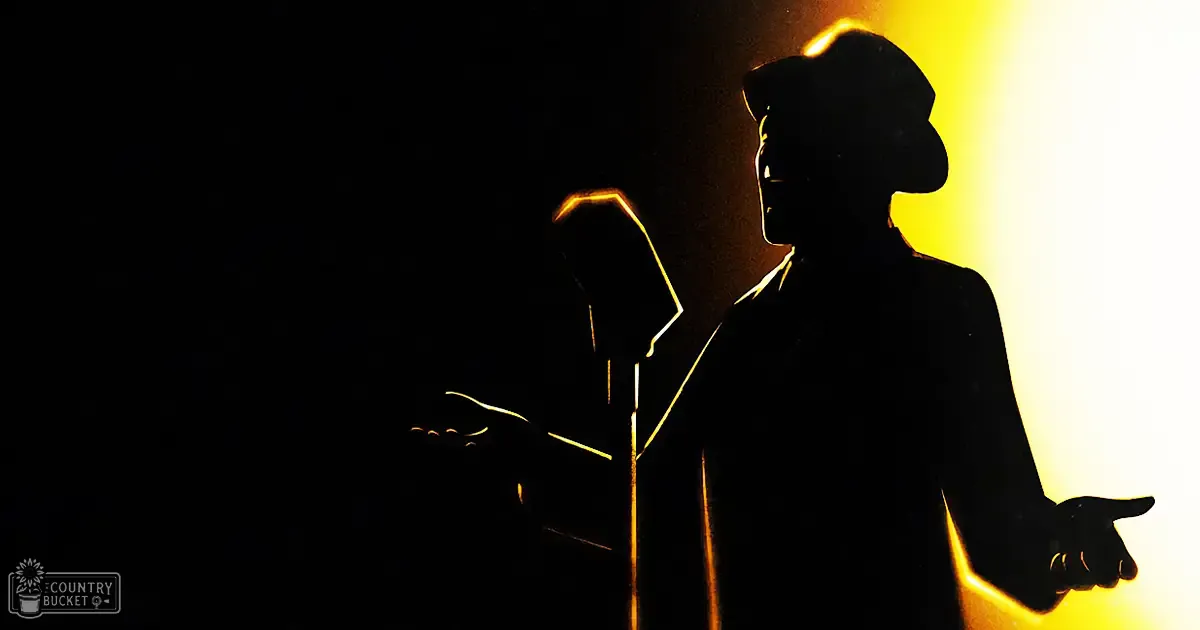 A male adult performer at a concert or stage event, wearing a dark jacket and wide-brimmed hat, silhouetted against dramatic stage lighting with bright yellow accents in an indoor venue during the evening.