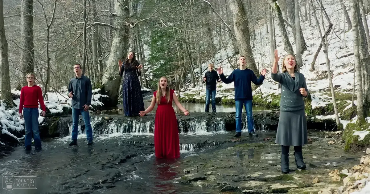 A group of adults and children singing in a snowy forest with snow-covered trees and rocks, during winter daytime.