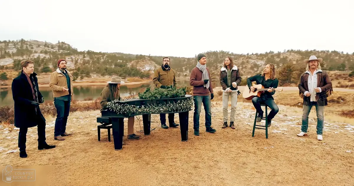 Musical performance in a scenic outdoor setting near a body of water with diffused light suggesting late afternoon or early evening; earthy tones dominate the scene.