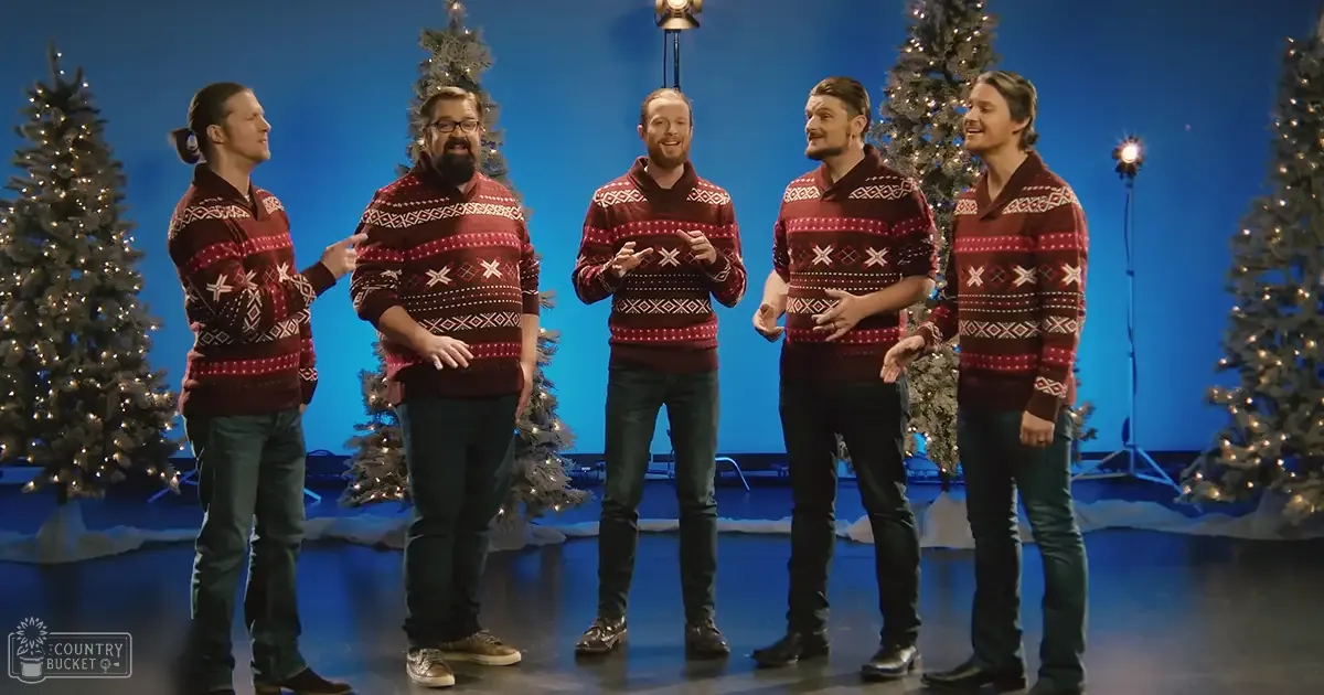 A group of young adult males performing in front of Christmas trees at an indoor event or studio setting with a blue backdrop.