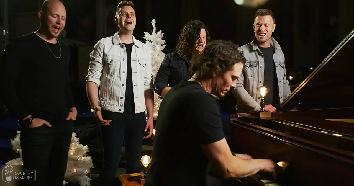 An adult male singer playing piano with four other singers in an indoor performance space, decorated for Christmas and illuminated by artificial lighting.