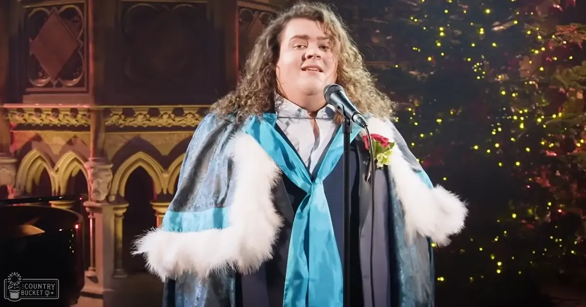 Adult male in a modern coat with fur trim, performing indoors near stained glass windows during the holiday season under artificial stage lighting.