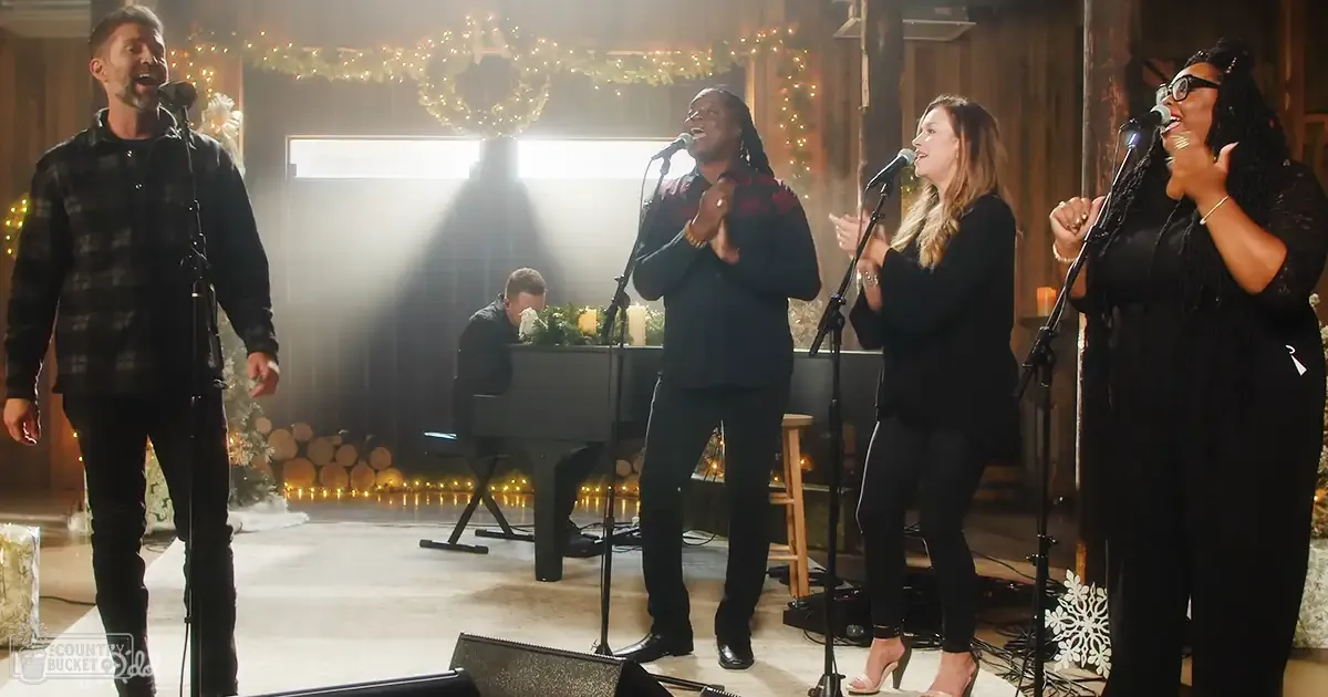 A group singing performance in an indoor Christmas-decorated setting with wooden walls, a grand piano, and festive lights.