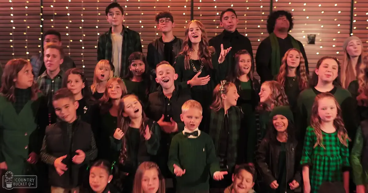 A group photo taken indoors, possibly in a school or community setting with children wearing dark green garments and string lights on the wall suggesting it might be around Christmas time.