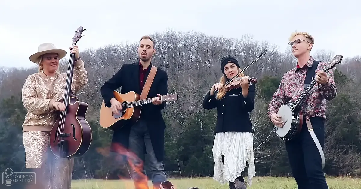 Adult female with wide-brimmed beige hat plays large upright bass; adult male in dark suit jacket strums acoustic guitar; adult female in black top and white skirt holds violin; adult male in patterned shirt plays banjo. Open field, bare trees suggest autumn or winter late afternoon light.