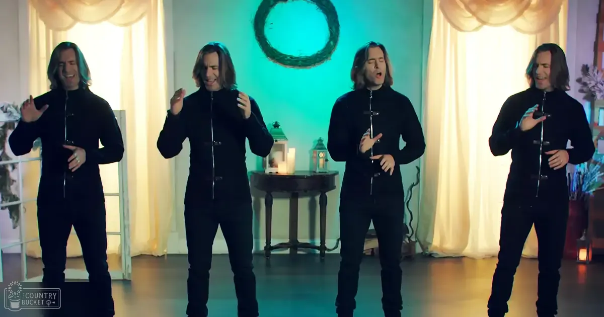 Adult male performers in a festive indoor setting, wearing black zip-up uniforms with high necklines and small chest devices. Curtains, a wreath, and decorative elements suggest a winter or Christmas theme.