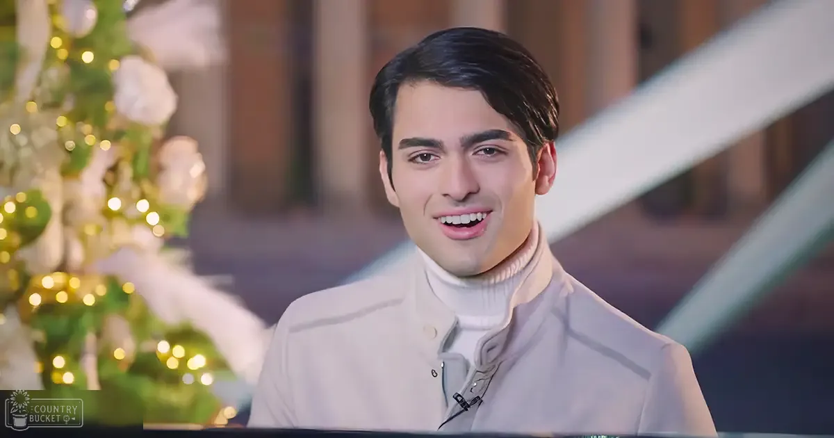 A young adult male wearing a light-colored, relaxed-fit jacket over a white turtleneck sweater stands indoors amidst festive decorations with artificial lighting.