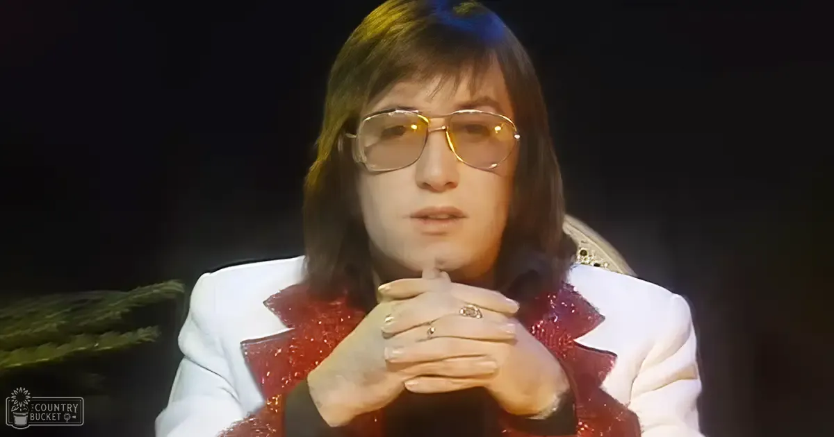 Young adult male in a relaxed fit, white blazer with red sequined detailing, seated indoors in a professional or studio environment, wearing gold-rimmed glasses and multiple rings.