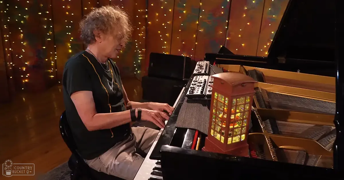 A middle-aged adult male plays a grand piano in an indoor setting, surrounded by home decor like string lights and a decorative lantern.