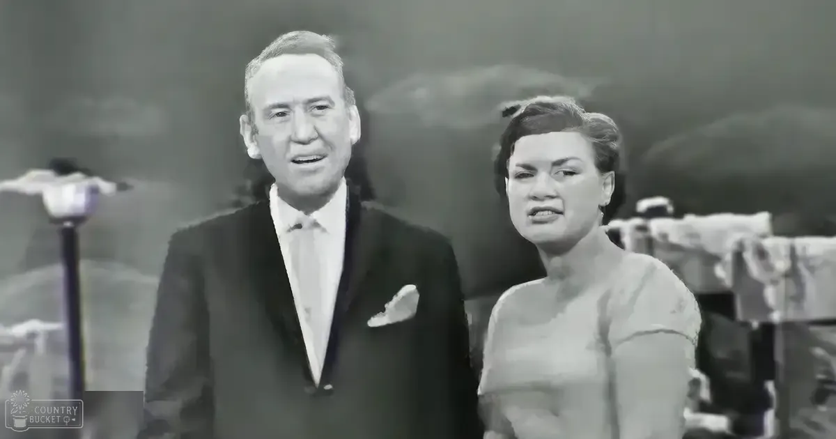 A middle-aged man wearing a dark suit jacket, white shirt, and light-colored pocket square stands on stage with a woman in an adult years' dress beside him; the setting suggests a performance venue.