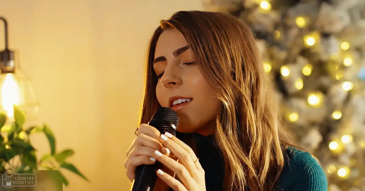A young adult female singing into a microphone in an indoor setting decorated for Christmas, with warm indoor lighting and a visible teal sweater.