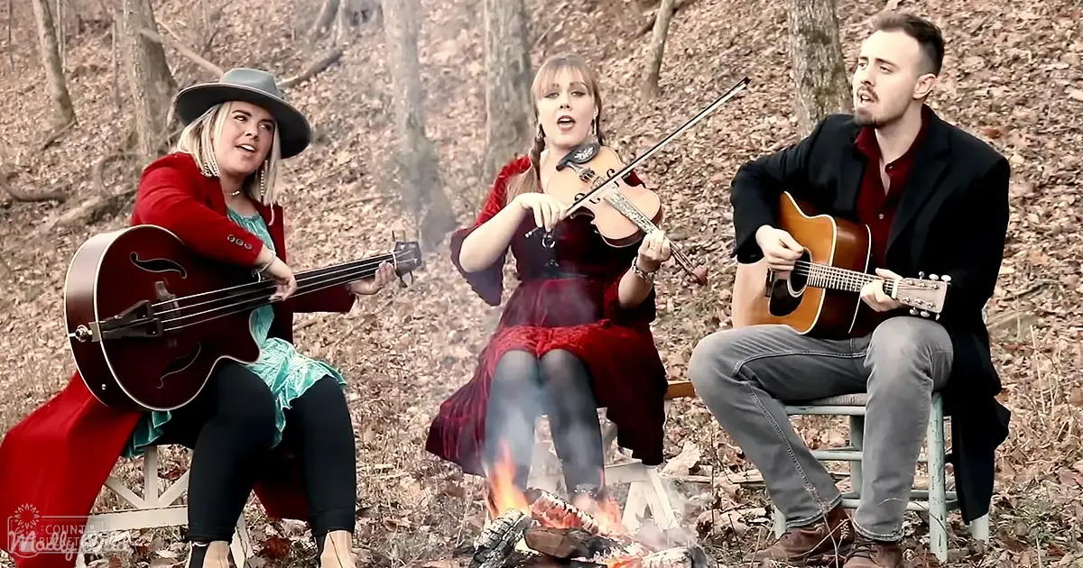 A group of musicians in a wooded outdoor setting with fallen leaves and simple wooden chairs. The scene suggests autumn or winter, with soft daylight filtering through the trees.