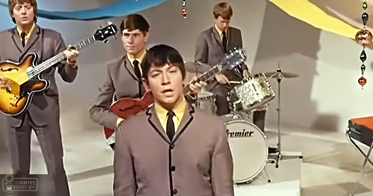 A young adult male singer in a gray suit jacket and yellow shirt performs on stage alongside other musicians playing instruments like the guitar, violin, and drums, set against light-colored studio walls with decorative fabric.