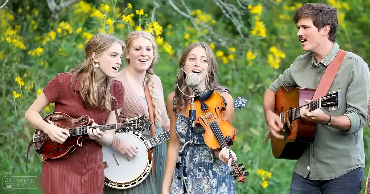 A group of young adult females performing music with string instruments in a lush outdoor setting with green foliage and yellow flowers on a bright sunny day.