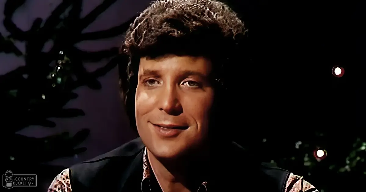 A young adult male wearing a dark, patterned shirt with leopard print on collar and cuffs stands indoors during what appears to be a festive season, possibly Christmas, surrounded by dimly lit decorative elements.