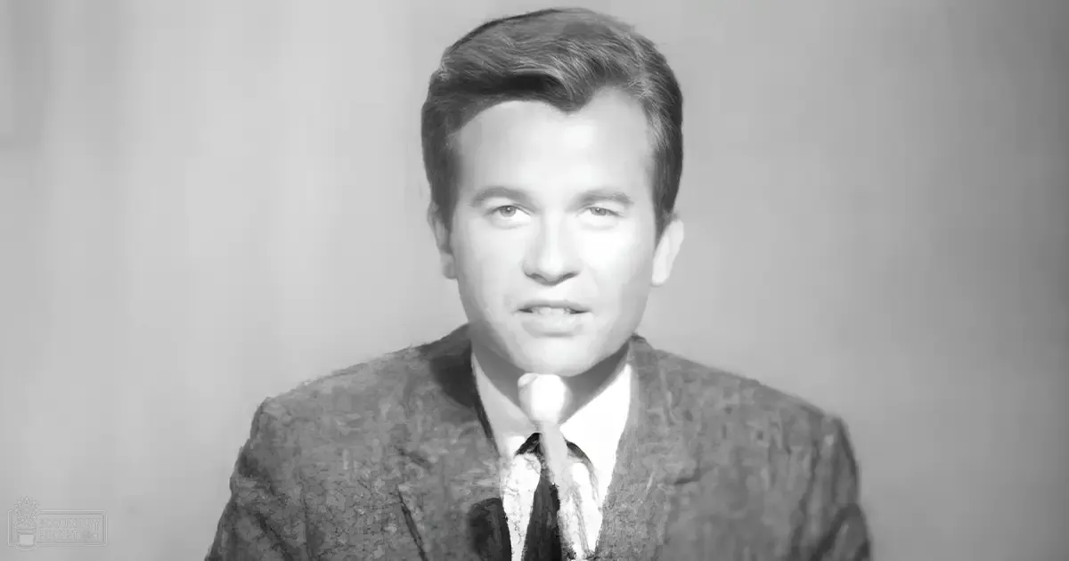 A young adult male in a studio setting, wearing a textured blazer over a collared shirt and tie, holding a microphone.