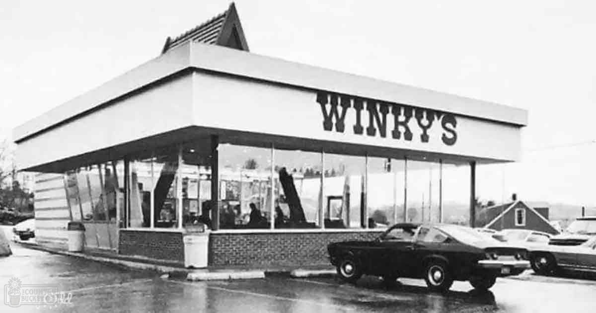 An urban street scene with Winky's diner, simple mid-20th-century furniture inside, parked cars and trees in the background with overcast daylight.