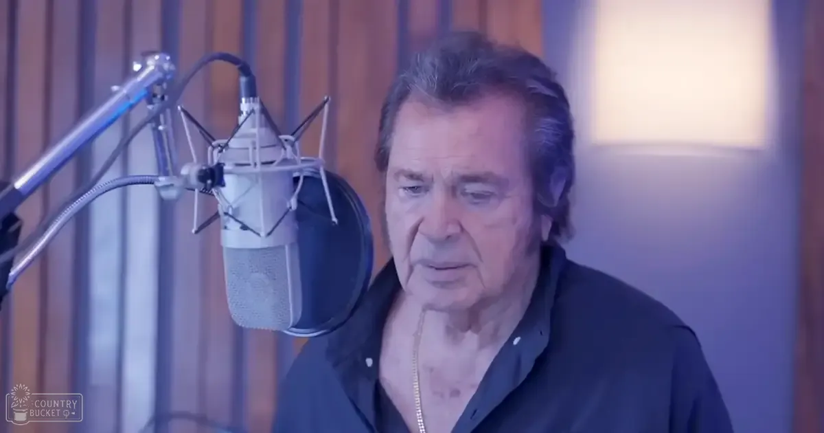 An older adult male in a recording studio with short graying hair wearing a dark collared shirt and thin chain necklace, surrounded by professional microphone equipment under soft diffused lighting.
