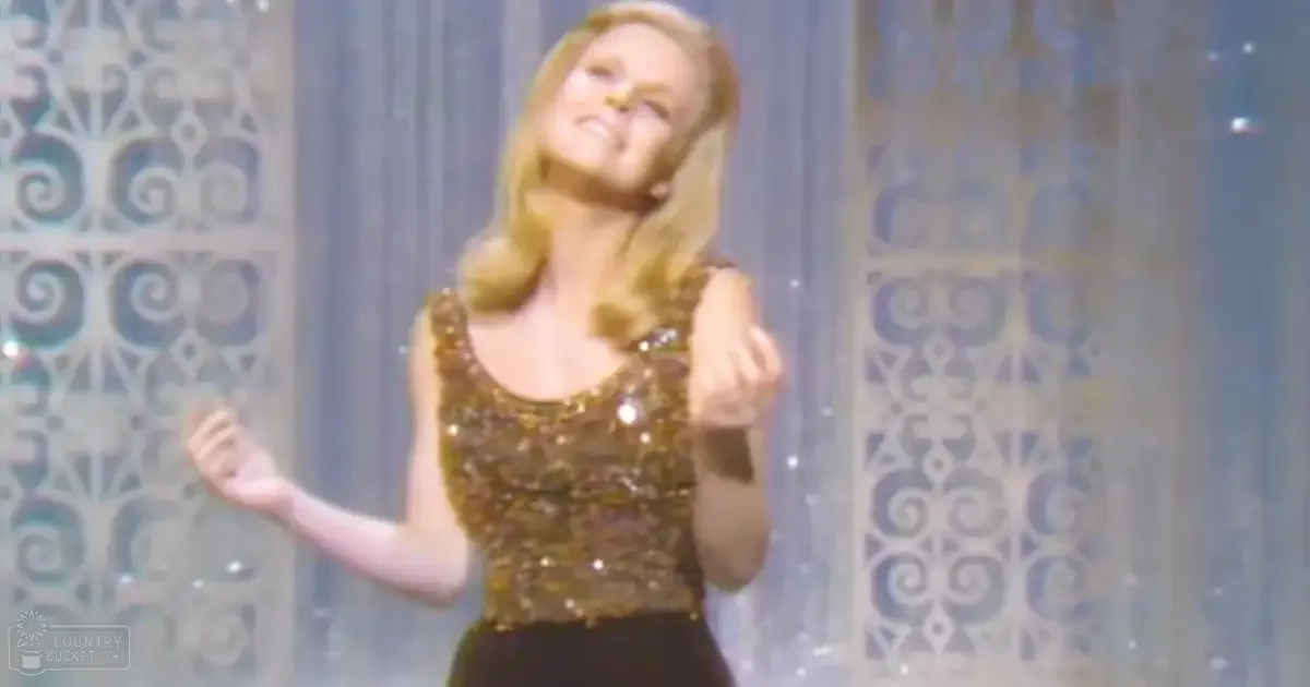A young adult female wearing a form-fitting, sleeveless sequined gold top with black pants stands on what appears to be a stage or performance area with decorative metalwork and sheer curtains behind her.