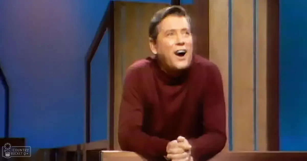 A middle-aged adult male wearing a maroon turtleneck sweater in an indoor setting with blue walls and wooden paneling, possibly a studio or stage.