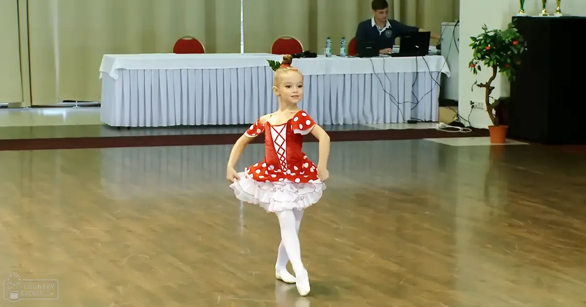 A young child in a red and white polka dot ballet tutu stands in an indoor dance studio with long tables, chairs, and a man using a laptop.