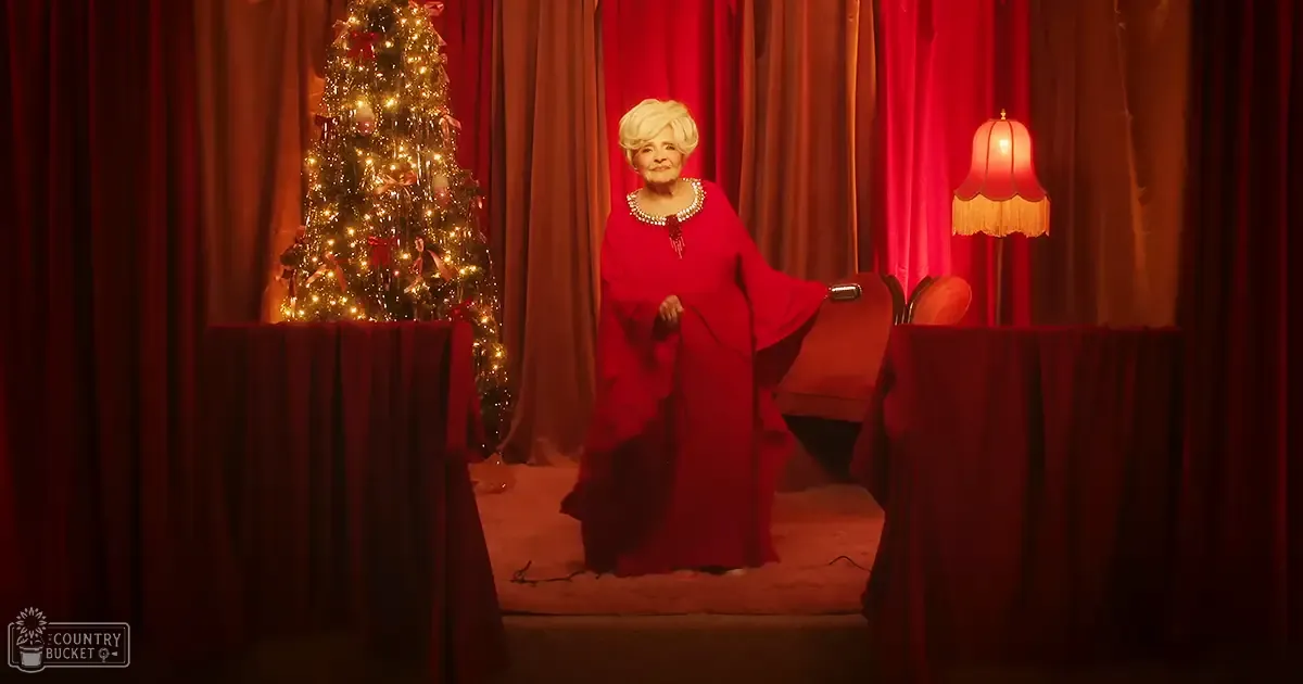 An older adult female wearing a red long-sleeved dress with loose, flowing fit and deep V-neck, accessorized with pearl necklace and earrings, stands in front of a Christmas tree decorated for the holiday season.